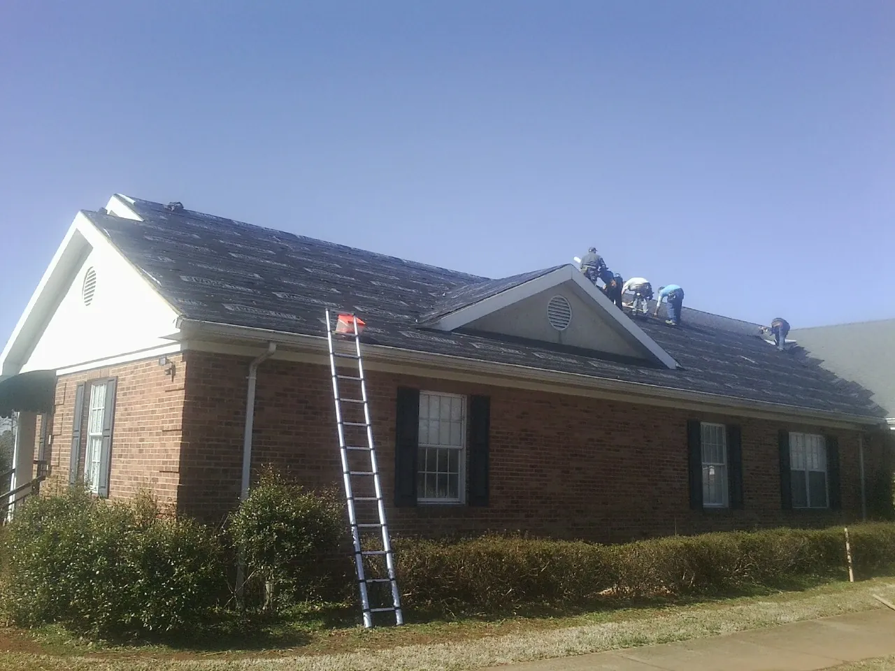 Crew tearing shingles off a brick ranch roof with a ladder set — Metal Roof Repair work by our crew in Garrison